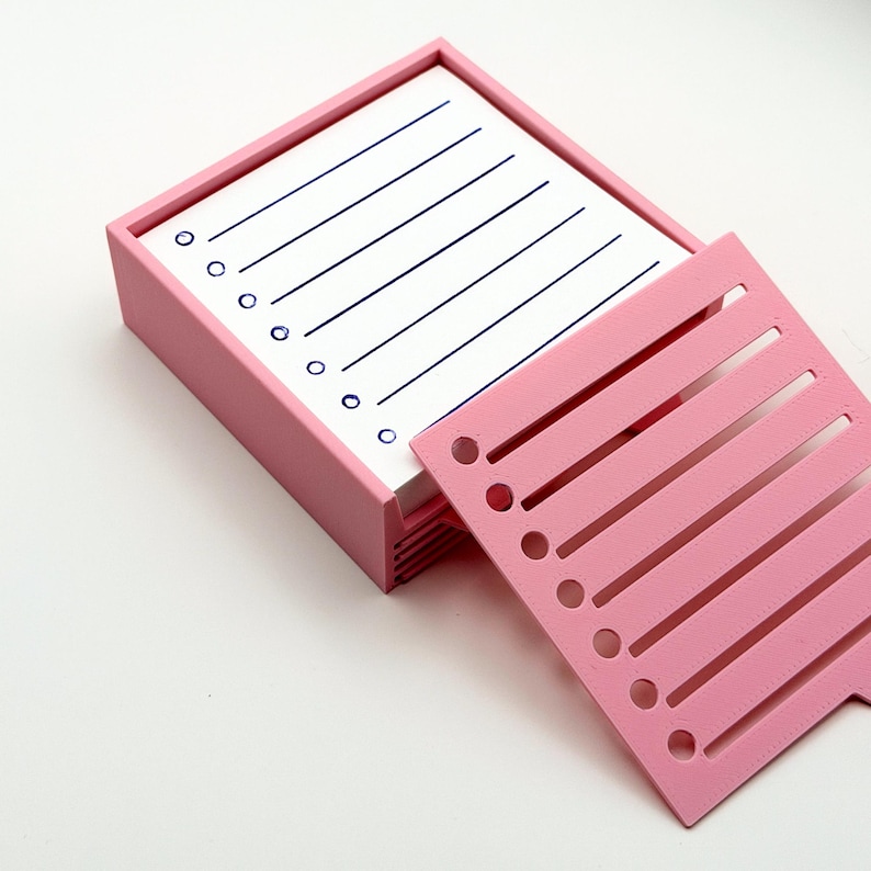 May include: A pink desk organizer with a stack of lined white notepaper and a pink divider. The notepaper has blue lines and circles. The divider has slots and holes. The organizer is on a white surface.