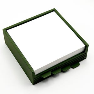 May include: A square, olive green desk organizer with a stack of white notepaper. The organizer has a rectangular frame and a series of angled tabs on the front. The design is simple and modern, suitable for office or home use.