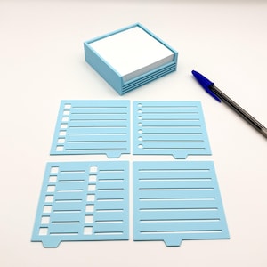 May include: A light blue desk organizer with a stack of white notepaper. Four light blue stencils with lined and dotted designs are arranged below the organizer. A blue and black pen rests on the surface.