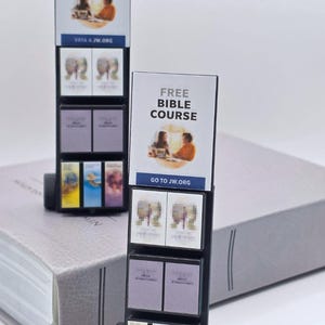May include: Miniature JW Literature cart with the text "JW Literature cart". The cart has multiple shelves with small books and signs that read "FREE BIBLE COURSE" and "CLASES DE LA BIBLIA GRATIS". The cart is black with wheels.
