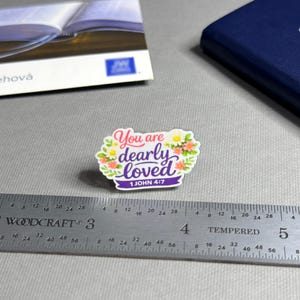 May include: A colorful sticker with the text "You are dearly loved" and the Bible verse 1 John 4:7. The sticker is surrounded by a floral design with pink, yellow, and white flowers. A ruler is next to the sticker, measuring in inches.