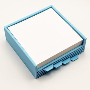 May include: A light blue square notepad holder with a stack of white notepaper. The holder has a frame around the top and a series of tabs on the side. The tabs are also light blue.