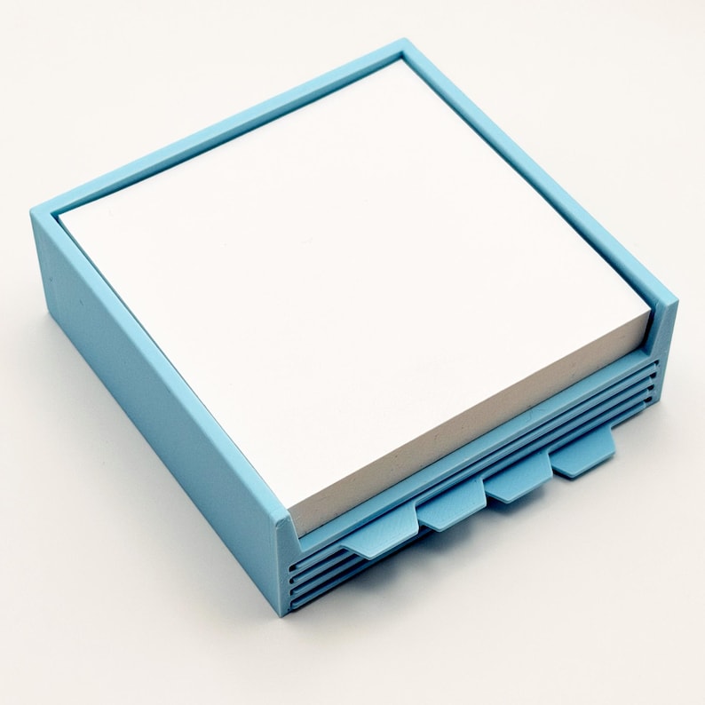 May include: A light blue square notepad holder with a stack of white notepaper. The holder has a frame around the top and a series of tabs on the side. The tabs are also light blue.