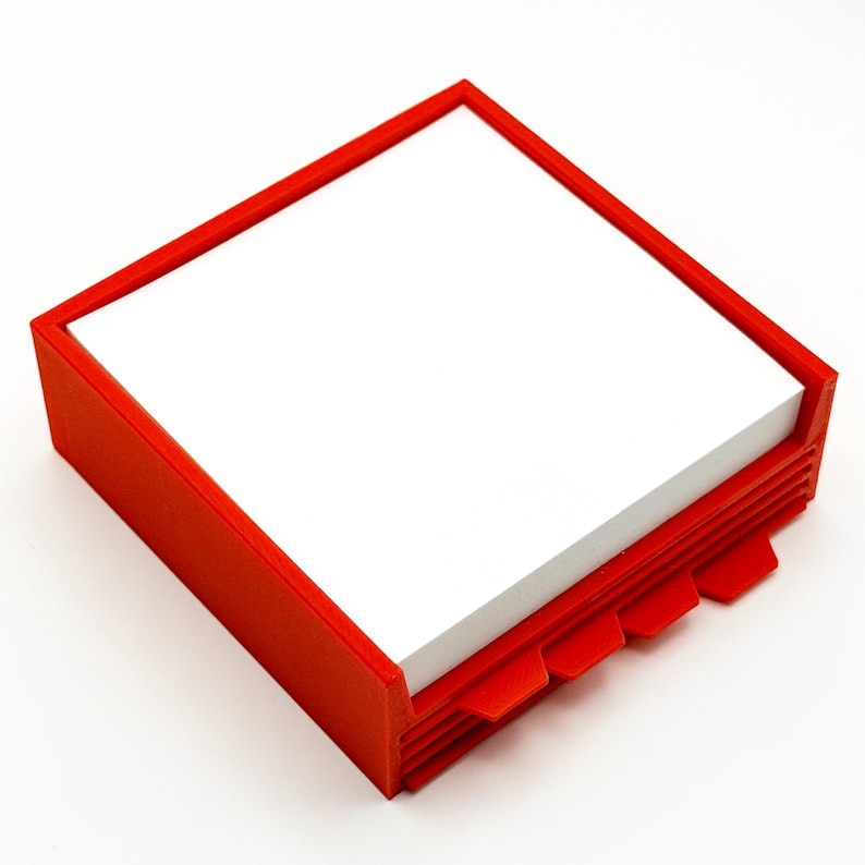May include: A red square notepad holder with a stack of white paper. The holder has a rectangular frame and a series of tabs on the bottom edge. The design is simple and modern.