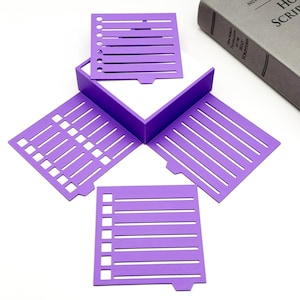 May include: A purple papercraft box with four open sides, each featuring a different design of cut-out lines and shapes. Several loose purple paper pieces with similar designs are scattered around the box. A gray book is in the background.