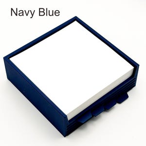 May include: A navy blue square notepad holder with a stack of white notepaper. The holder has a dark blue frame and a series of angled tabs on the bottom edge. The text "Navy Blue" is in the upper left corner.