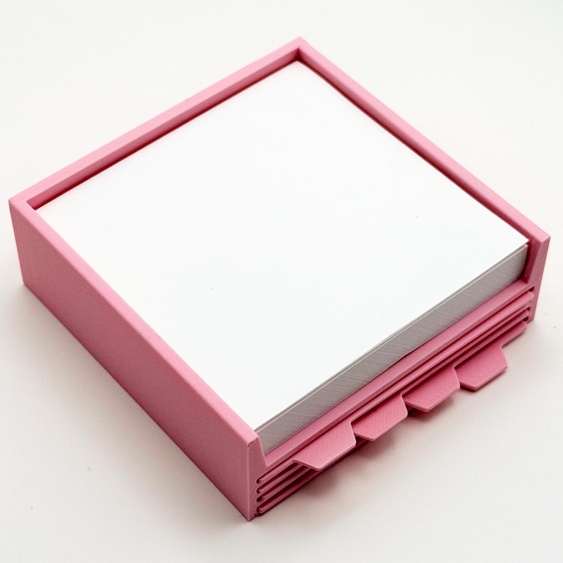 May include: A pink square notepad holder with a stack of white paper inside. The holder has a raised border and a series of pink tabs along one side. The tabs are used to separate the pages.