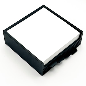 May include: A black square notepad holder with a stack of white notepaper. The holder has a simple, modern design with a frame around the top and a series of tabs on the side for easy access to the paper.
