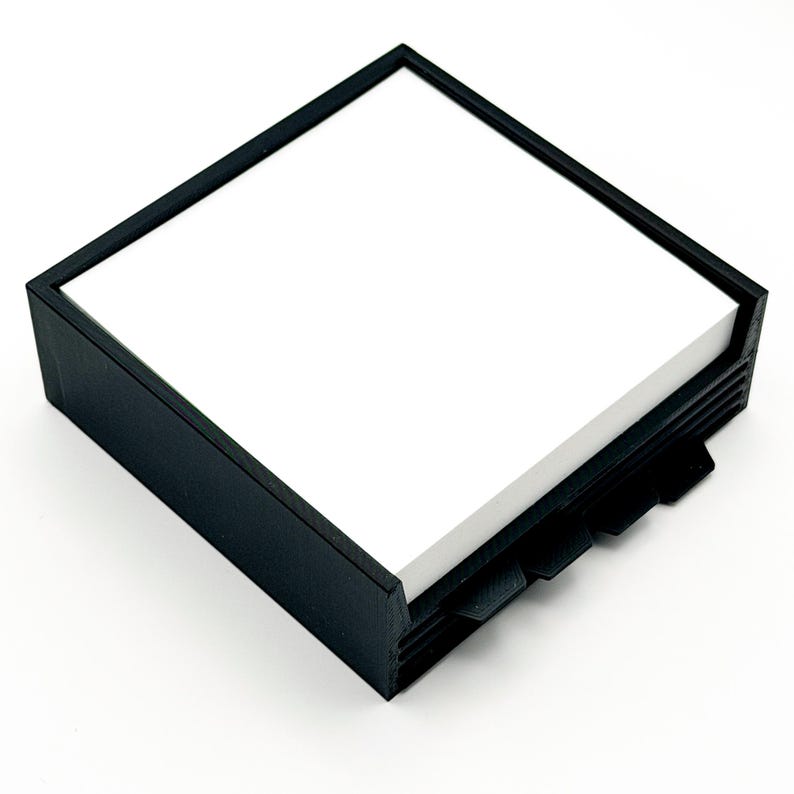 May include: A black square notepad holder with a stack of white notepaper. The holder has a simple, modern design with a frame around the top and a series of tabs on the side for easy access to the paper.