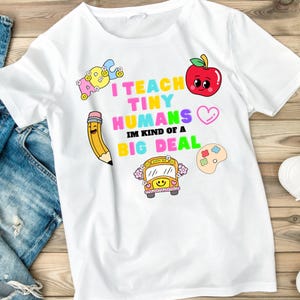 Puede incluir: Camiseta blanca con un diseño gráfico colorido. El diseño incluye el texto "I TEACH TINY HUMANS IM KIND OF A BIG DEAL" con ilustraciones de una manzana, un lápiz, un autobús escolar y una paleta de pintura. La camiseta es de material suave.