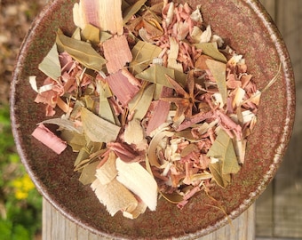 May include: A brown ceramic bowl filled with a mixture of dried herbs and wood chips. The mixture includes light brown wood chips, pink wood chips, and green leaves.