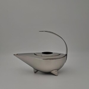 May include: A silver-colored teapot with a curved handle and three small feet. The teapot has a spout and a circular opening on top, and a lid. The design is modern and minimalist.