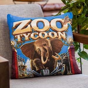 May include: A blue decorative pillow featuring the text "ZOO TYCOON" in gold lettering. The pillow has an illustration of an elephant, a tiger, and a giraffe. The pillow is resting on a gray chair.