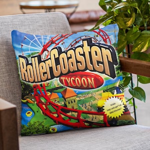 May include: A square pillow featuring the RollerCoaster Tycoon game cover art. The design includes a red roller coaster, a Ferris wheel, and the game's logo in gold and red. The pillow is on a gray chair.