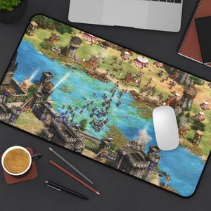以下が含まれることがあります： 中世の戦闘シーンのカラフルなイラストが描かれた長方形のマウスパッド。川、建物、兵士が描かれています。白いコンピューターマウスがパッドの上にあります。