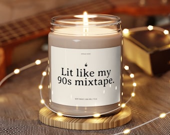 Op de afbeelding: Een bruine sojawas kaars met een wit label waarop staat "Lit like my 90s mixtape." De kaars staat op een houten onderzetter en is omringd door witte lichtsnoeren.
