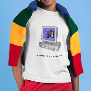 Puede incluir: Una camiseta blanca con un gráfico de una pantalla de ordenador con el texto "RUNNING ON DIAL-UP" debajo. La persona lleva pantalones cortos rojos y una bufanda multicolor.