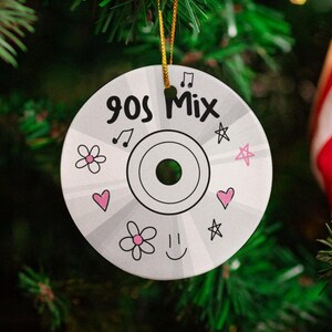 May include: A round, silver ornament with the text "90s Mix" and decorative doodles. The ornament features musical notes, stars, flowers, and hearts in pink and black. A gold string is attached for hanging on a Christmas tree.