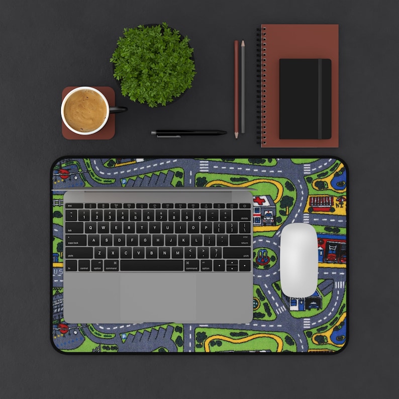 Mousepad featuring the iconic 90s and 2000s city street carpet design, with colorful illustrated roads, buildings, and vehicles. Styled on a black desk with a laptop, coffee cup, notebooks, and pencils.
