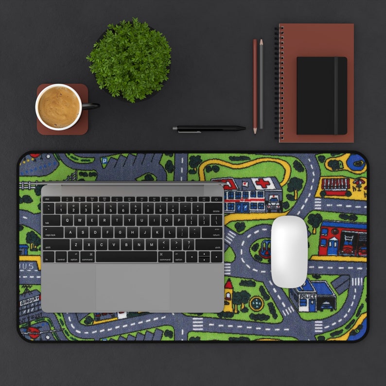 Mousepad featuring the iconic 90s and 2000s city street carpet design, with colorful illustrated roads, buildings, and vehicles. Styled on a black desk with a laptop, coffee cup, notebooks, and pencils.