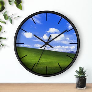 May include: Circular wall clock with a black frame. The clock face displays a vivid image of a green hill beneath a blue sky with white clouds. Black hands and hour markers complete the design.