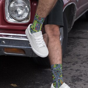 May include: A pair of white canvas sneakers with colorful socks featuring a city street design. The socks are green, blue, and yellow. The sneakers are worn by a person sitting on the hood of a red car.
