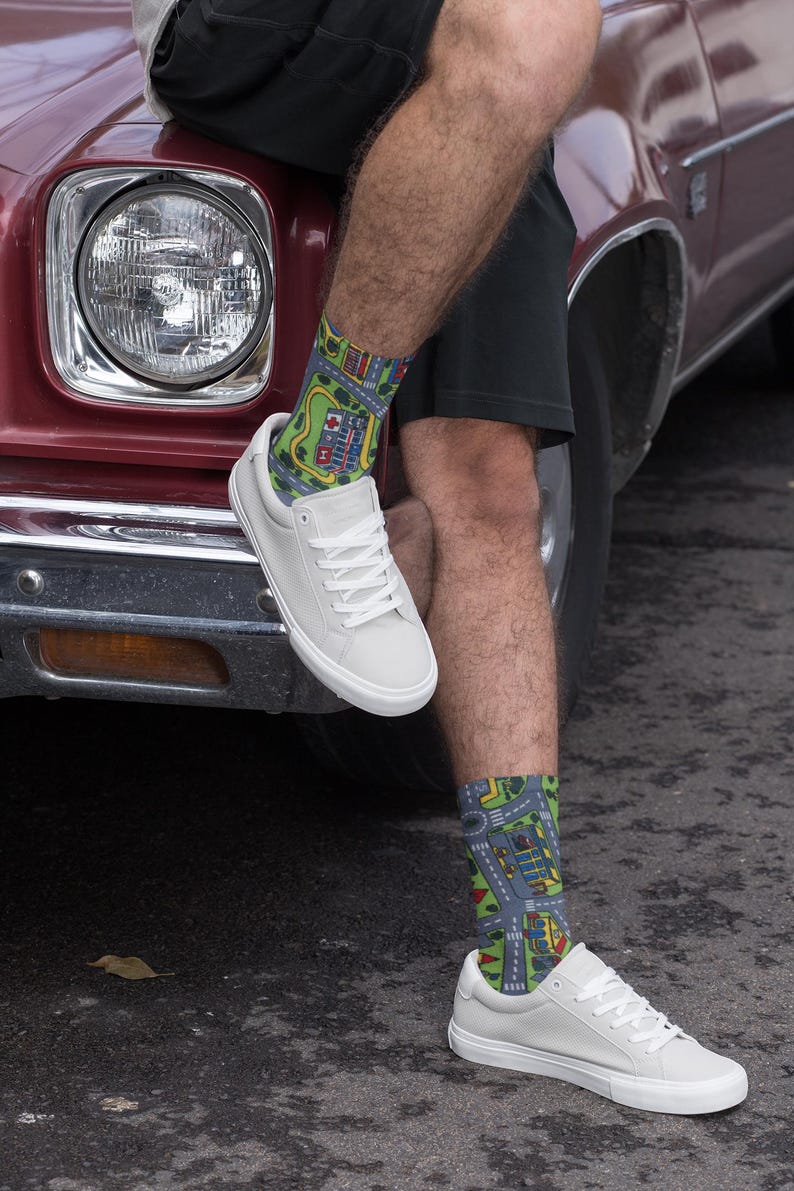 May include: A pair of white canvas sneakers with colorful socks featuring a city street design. The socks are green, blue, and yellow. The sneakers are worn by a person sitting on the hood of a red car.