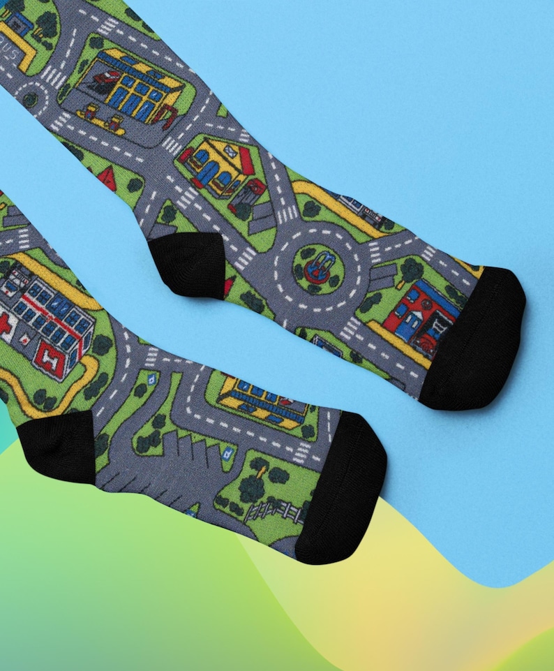 May include: A pair of black socks with a colorful playmat design. The socks feature a gray road with yellow lines, green grass, and colorful buildings.