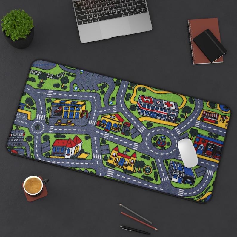 Close-up of mousepad featuring the iconic 90s and 2000s city street carpet design, with colorful illustrated roads, buildings, and vehicles. Styled on a black desk with a laptop, coffee cup, notebooks, and pencils.