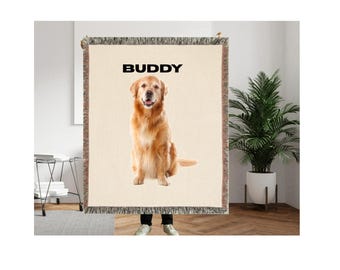 May include: A beige woven throw blanket featuring a golden retriever dog with the name "BUDDY" in black block letters. The dog is sitting and looking forward. The blanket has a fringed edge.