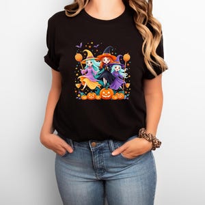 May include: Black t-shirt featuring a colorful Halloween graphic. The design shows three cartoon witches with orange balloons, surrounded by pumpkins, stars, and bats. The witches wear pointed hats and dresses in purple, orange, and teal.