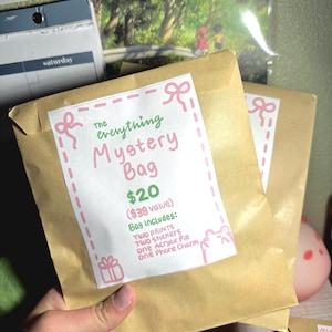 Puede incluir: Dos sobres de papel marrón con texto rosa y verde que dice "The everything Mystery Bag 20 € (38 € de valor) La bolsa incluye: DOS IMPRESIONES Dos pegatinas un pin acrílico un charm de teléfono"