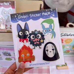 May include: A sticker pack featuring characters from Studio Ghibli films. The pack includes a black cat with a red bow, a black and pink creature with a star, a blue cat with a leaf, a fire spirit, and a No Face character.