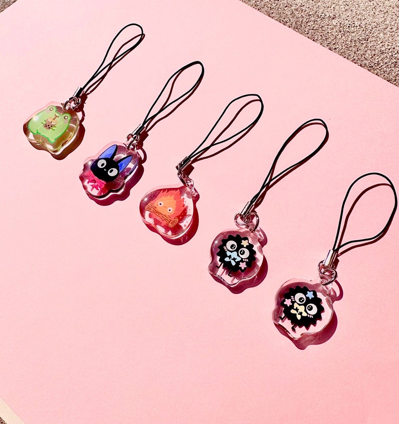 May include: Five colorful phone charms with different characters. The charms are attached to black strings. The charms are on a pink background.