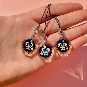 May include: Three black and white cartoon character charms with pink, blue, and yellow accents. The charms are attached to black string phone straps.