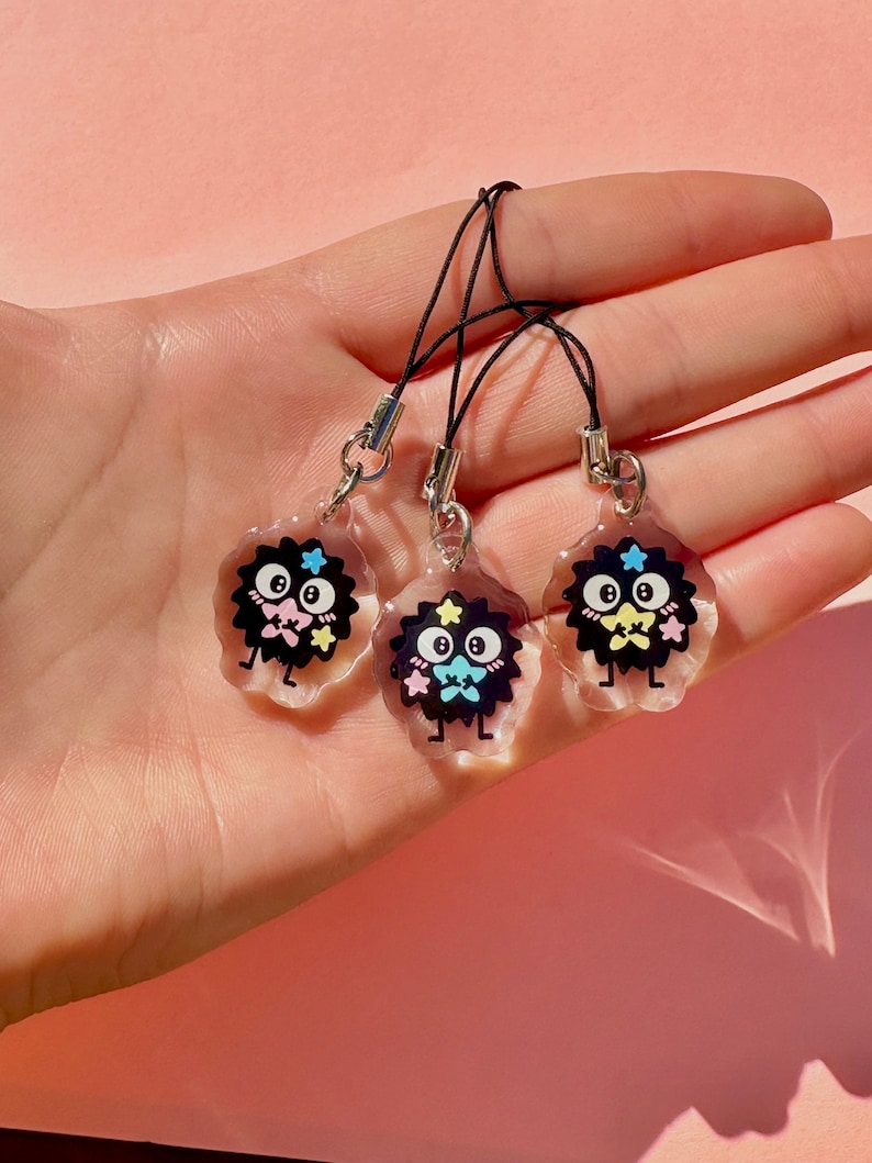 May include: Three black and white cartoon character charms with pink, blue, and yellow accents. The charms are attached to black string phone straps.
