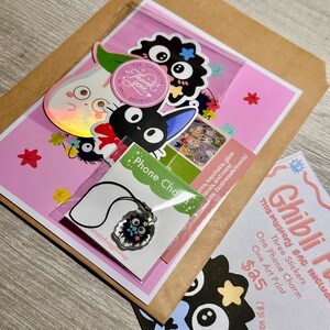 May include: A mystery bag with Ghibli-themed items, including stickers, a phone charm, and an art print. The bag is packaged in a pink and brown envelope, with a variety of colorful stickers and a necklace with a charm. The text "Thank you" is visible on a round sticker.