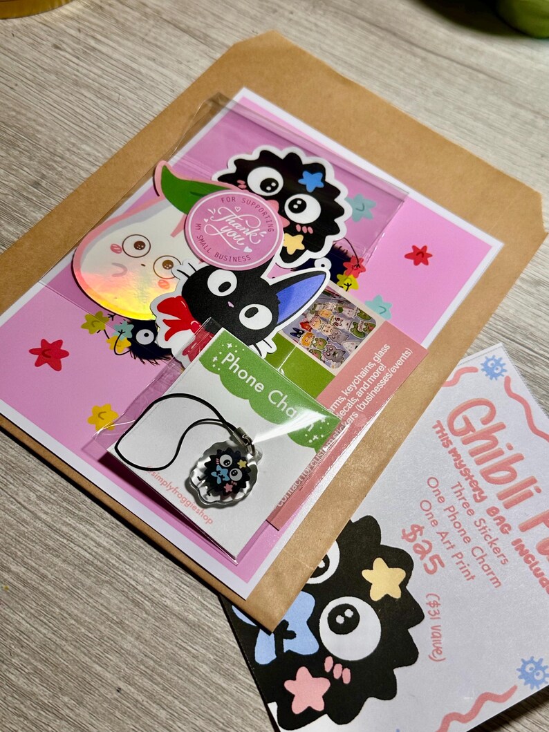 May include: A mystery bag with Ghibli-themed items, including stickers, a phone charm, and an art print. The bag is packaged in a pink and brown envelope, with a variety of colorful stickers and a necklace with a charm. The text "Thank you" is visible on a round sticker.