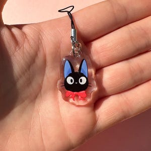 May include: A clear charm with a black cat with blue ears and a red bow. The charm is attached to a black string.