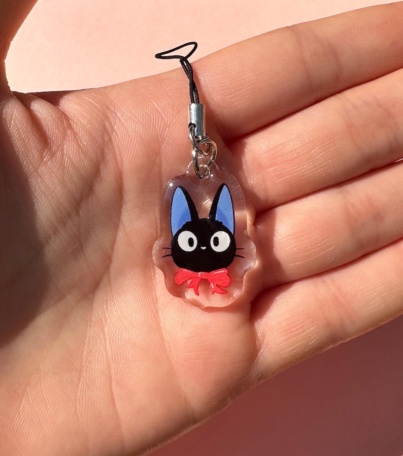 May include: A clear charm with a black cat with blue ears and a red bow. The charm is attached to a black string.