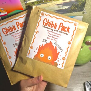 K&ouml;nnte beinhalten: Zwei braune Papiert&uuml;ten mit der Aufschrift "Ghibli Pack" und einer Cartoon-Feuerfigur. Jede T&uuml;te enth&auml;lt einen Telefonanh&auml;nger, einen Acryl-Pin, Aufkleber und einen Kunstdruck.