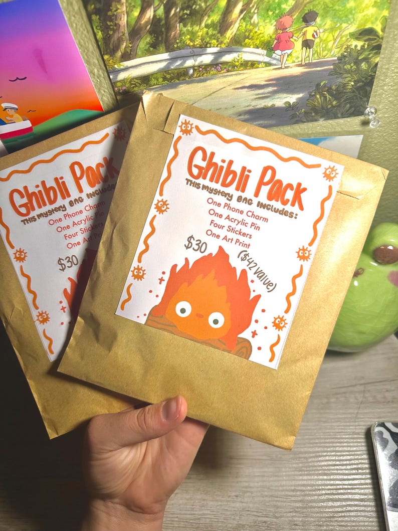 K&ouml;nnte beinhalten: Zwei braune Papiert&uuml;ten mit der Aufschrift "Ghibli Pack" und einer Cartoon-Feuerfigur. Jede T&uuml;te enth&auml;lt einen Telefonanh&auml;nger, einen Acryl-Pin, Aufkleber und einen Kunstdruck.