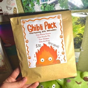 K&ouml;nnte beinhalten: Ein brauner Papiersack mit der Aufschrift "Ghibli Pack" mit orangefarbenem Text und einer Cartoon-Feuerfigur. Die Tasche enth&auml;lt einen Handy-Anh&auml;nger, einen Acryl-Pin, Aufkleber und einen Kunstdruck.