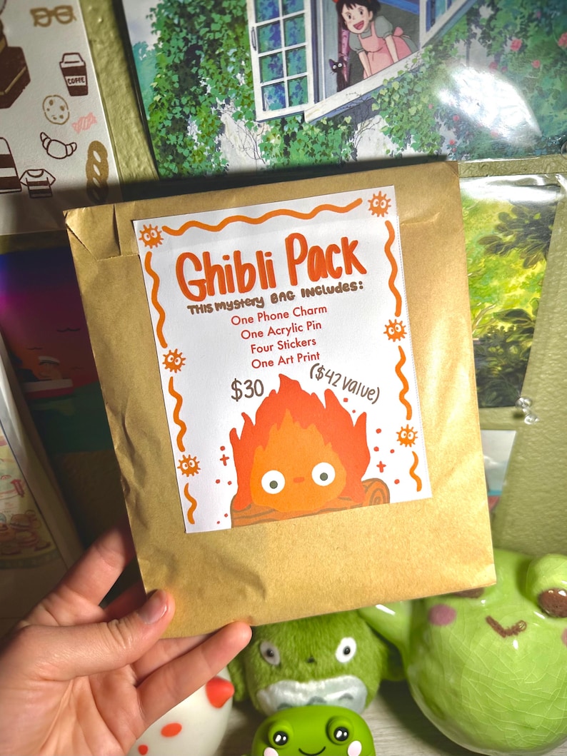 K&ouml;nnte beinhalten: Ein brauner Papiersack mit der Aufschrift "Ghibli Pack" mit orangefarbenem Text und einer Cartoon-Feuerfigur. Die Tasche enth&auml;lt einen Handy-Anh&auml;nger, einen Acryl-Pin, Aufkleber und einen Kunstdruck.