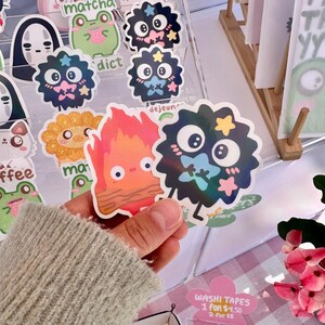 May include: A collection of colorful stickers featuring cartoon characters, including a fire spirit, a black and white creature with stars, and a green frog. The stickers are displayed on a clear acrylic shelf with the text "matcha", "dict", and "dejeun" visible.