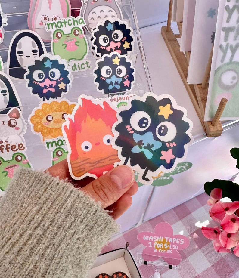 May include: A collection of colorful stickers featuring cartoon characters, including a fire spirit, a black and white creature with stars, and a green frog. The stickers are displayed on a clear acrylic shelf with the text "matcha", "dict", and "dejeun" visible.