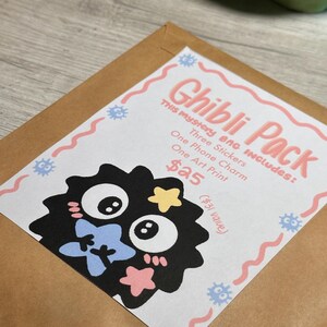 May include: A brown paper envelope with a white label featuring the text "Ghibli Pack." The label includes a black cartoon character with blue, yellow, and pink accents. The label also lists the contents: three stickers, one phone charm, and one art print.