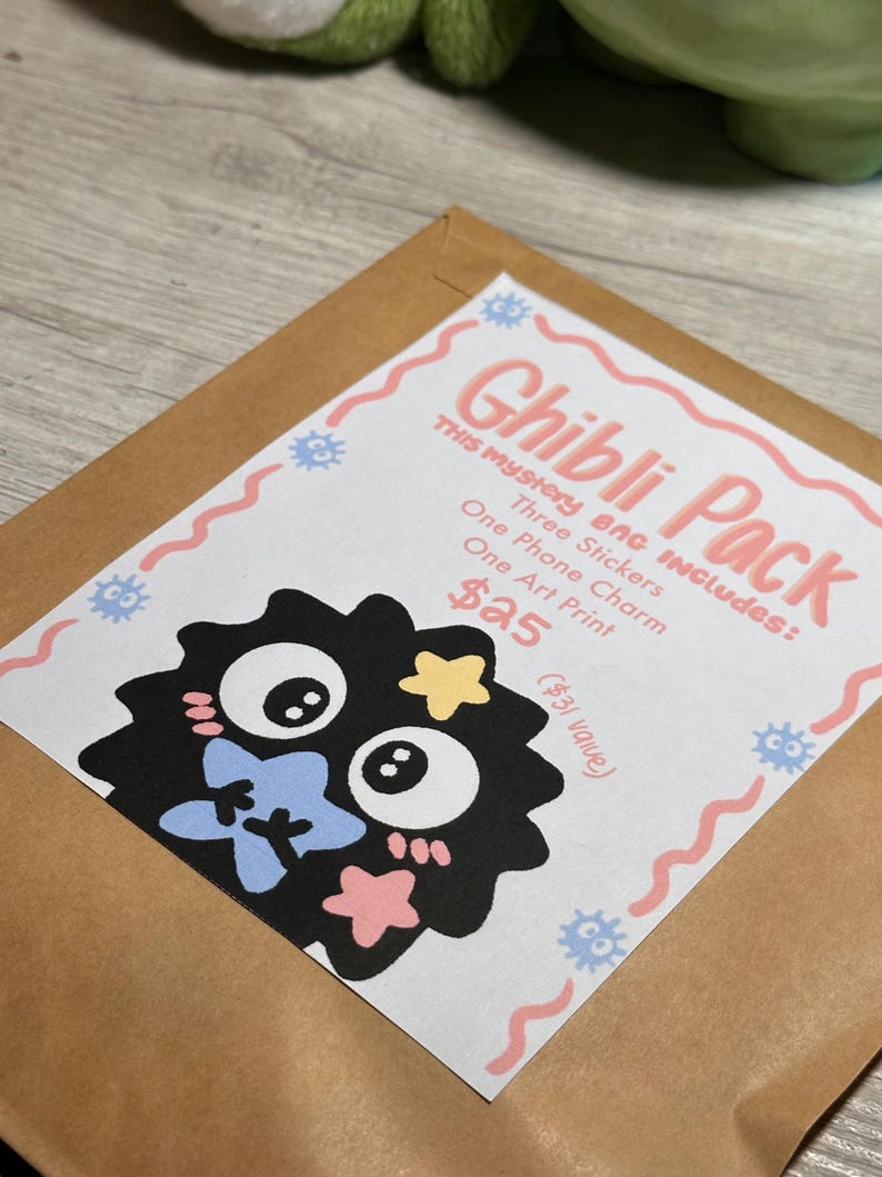 May include: A brown paper envelope with a white label featuring the text "Ghibli Pack." The label includes a black cartoon character with blue, yellow, and pink accents. The label also lists the contents: three stickers, one phone charm, and one art print.
