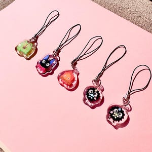 May include: Five clear acrylic charms with black straps, featuring cartoon characters. The charms include a green frog, a black cat with a pink bow, a fiery creature, and two black soot sprites with stars and flowers. The charms are on a pink surface.