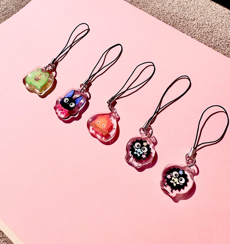 May include: Five clear acrylic charms with black straps, featuring cartoon characters. The charms include a green frog, a black cat with a pink bow, a fiery creature, and two black soot sprites with stars and flowers. The charms are on a pink surface.